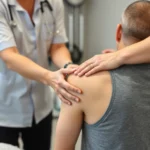 Physical therapist performing hands-on manual therapy and joint mobilization on patient's shoulder, clinical rehabilitation setting with modern equipment visible in background, professional healthcare environment