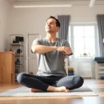 A patient in a bright physical therapy clinic performing a gentle stretching exercise while sitting cross-legged, eyes closed in peaceful concentration, with a physiotherapist observing nearby, natural window light streaming in