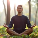 Person in peaceful meditation pose sitting cross-legged in serene nature environment, morning sunlight filtering through trees, calm expression, natural outdoor setting with soft green foliage
