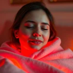 Person wrapped in warm red light therapy blanket in cozy bedroom during evening, soft ambient glow illuminating face with peaceful expression, comfortable home environment, photorealistic lighting