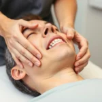 Physical therapist performing manual therapy on patient's jaw and temporomandibular joint, hands applying gentle mobilization technique, professional clinical setting with neutral background, patient in relaxed position