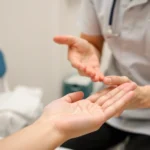 Professional hand therapist performing manual therapy on patient's palm and fingers, showing gentle mobilization technique with focused expression, medical clinic setting with neutral background