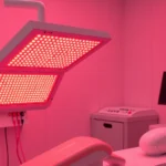 Professional red light therapy LED panel device emitting warm crimson light in modern clinical treatment room with stainless steel equipment and minimalist aesthetic, photorealistic