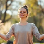 Person in peaceful meditation pose outdoors in natural sunlight, wearing neutral comfortable clothing, serene facial expression, soft focus background with blurred nature elements, photorealistic wellness imagery